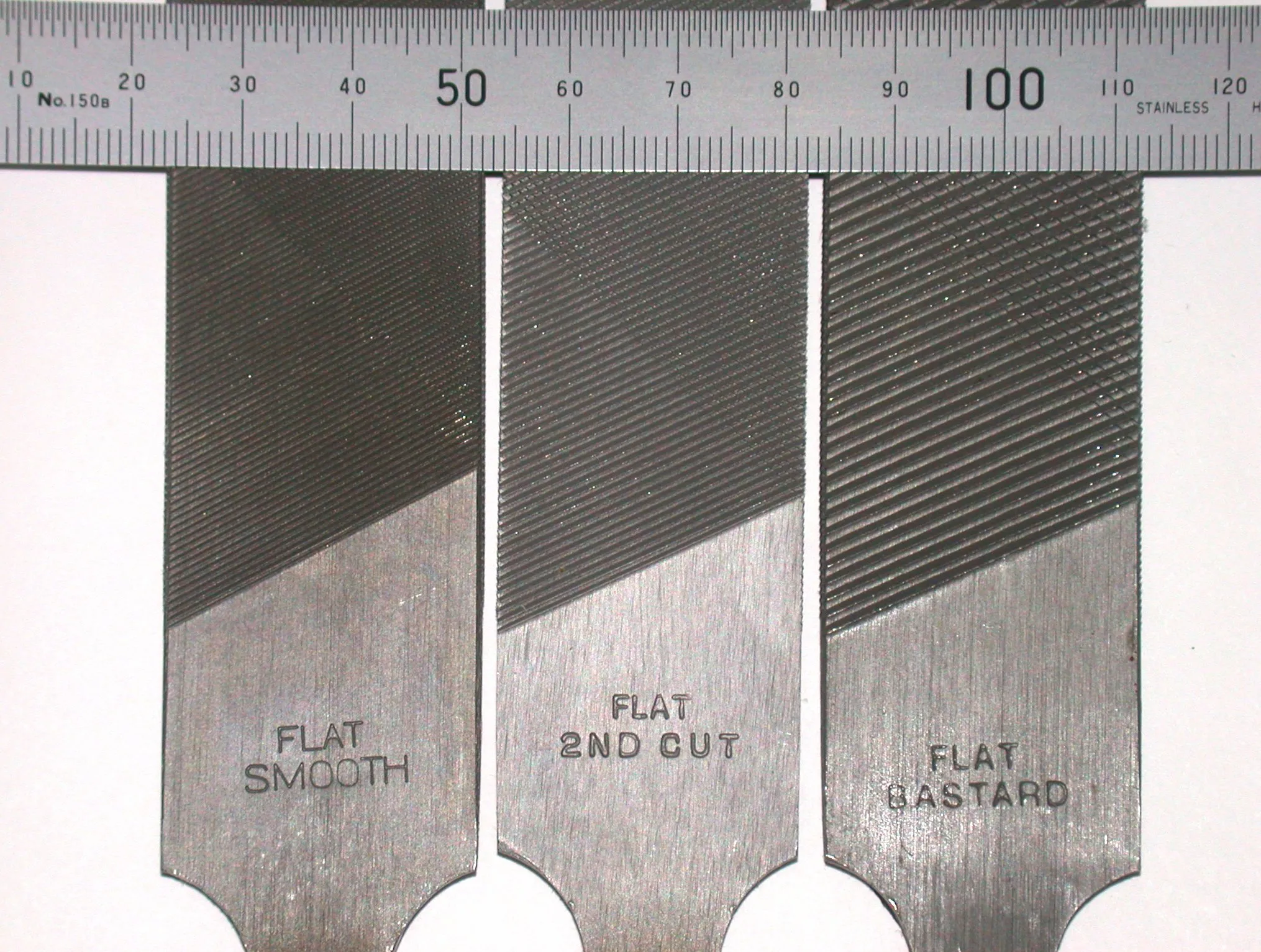 Três limas chatas de aço, lado a lado sob uma régua em milímetros, com as inscrições “Flat Smooth”, “Flat 2nd Cut” e “Flat Bastard” gravadas na espiga.