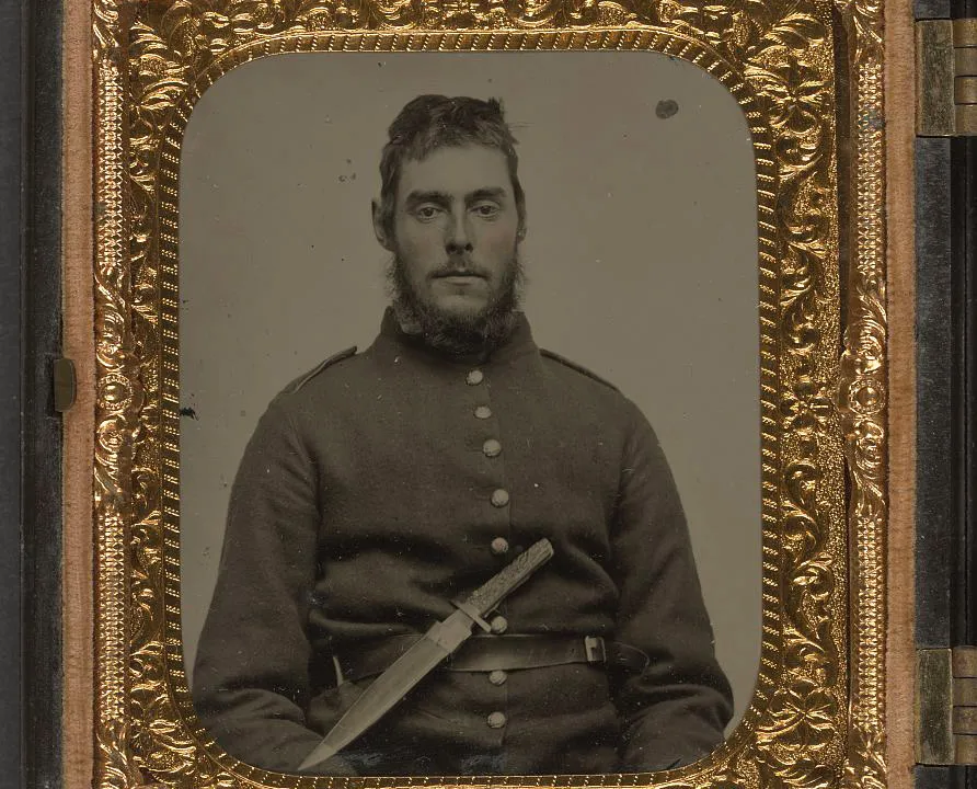 Ambrótipo colorido à mão de um soldado da União, entre 1861 e 1865, sentado de uniforme com uma faca bowie de lâmina larga presa ao cinto, a mão direita apoiada sobre o cabo.