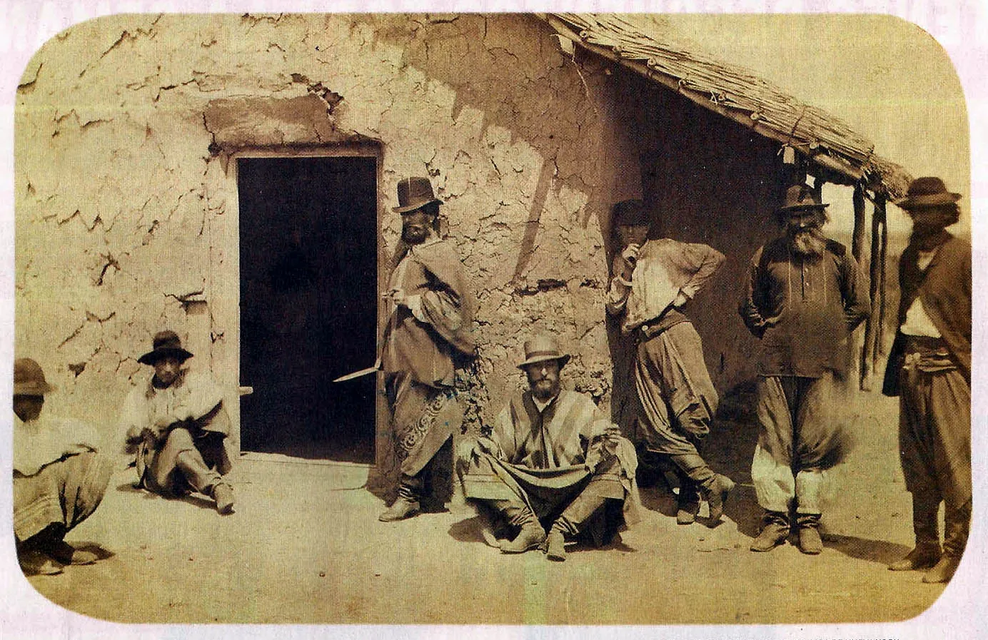 Fotografia em sépia, dos anos 1860, de seis gaúchos diante de um rancho de barro e palha; um deles, de pé junto à porta, segura uma faca longa; os outros sentados ou em pé, de chapéu, poncho e bombacha.