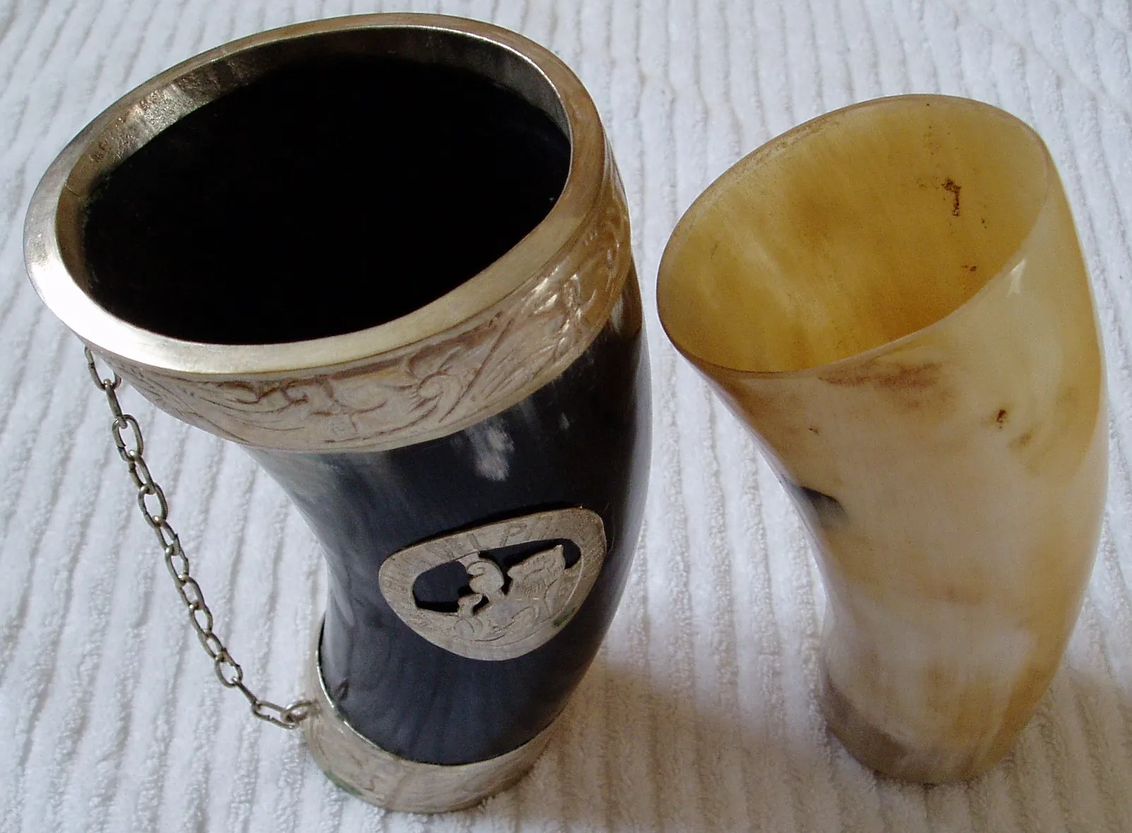 Duas guampas de chifre de boi sobre uma toalha branca: uma escura, quase preta, com aro e base de metal trabalhado e uma corrente; a outra clara, em tons de mel e branco, sem guarnição.