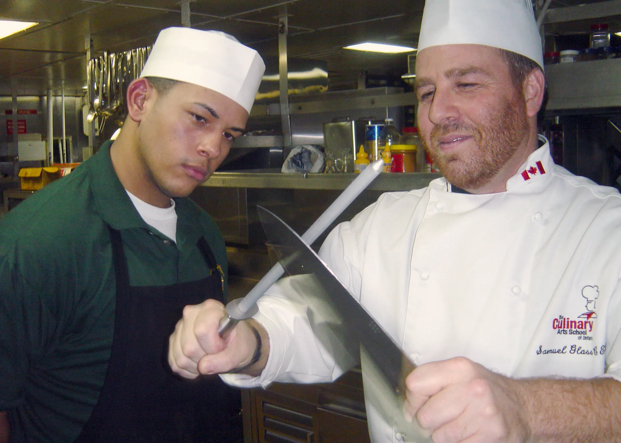Instrutor de cozinha demonstrando a passada da faca na chaira, com a haste na vertical e a lâmina descendo em ângulo