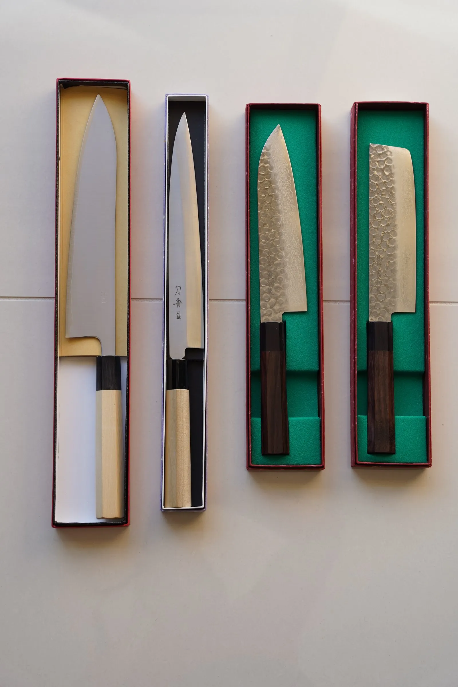 Quatro facas japonesas em caixas abertas, vistas de cima: um gyuto de cabo claro, uma yanagiba longa e estreita, um santoku de lâmina martelada e um nakiri retangular, os dois últimos com cabo escuro.