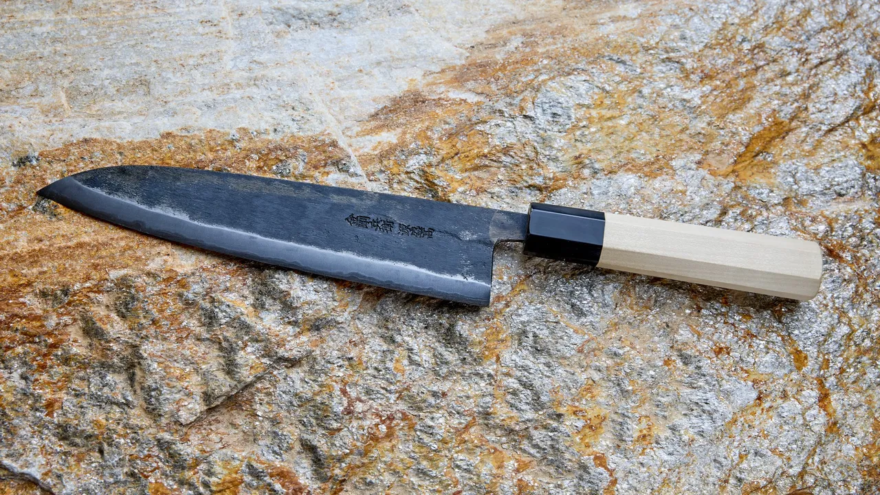 Faca de chef japonesa (gyuto) forjada à mão, com a lâmina escura de acabamento de forja e uma faixa polida junto ao fio, cabo octogonal de magnólia clara com virola preta, sobre uma pedra de tons cinza e ferrugem.
