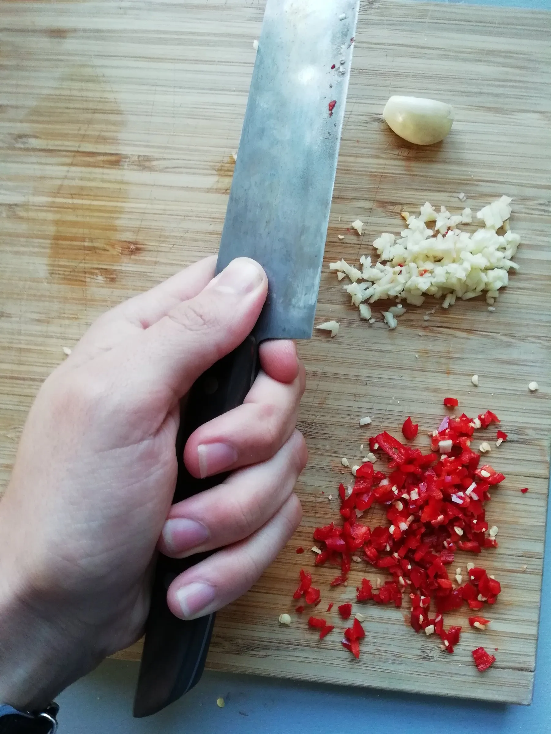 Mão segurando uma faca de cozinha em pegada de pinça sobre uma tábua de bambu, com o polegar e o indicador prendendo a lâmina logo à frente do cabo; ao lado, alho e pimenta vermelha picados.