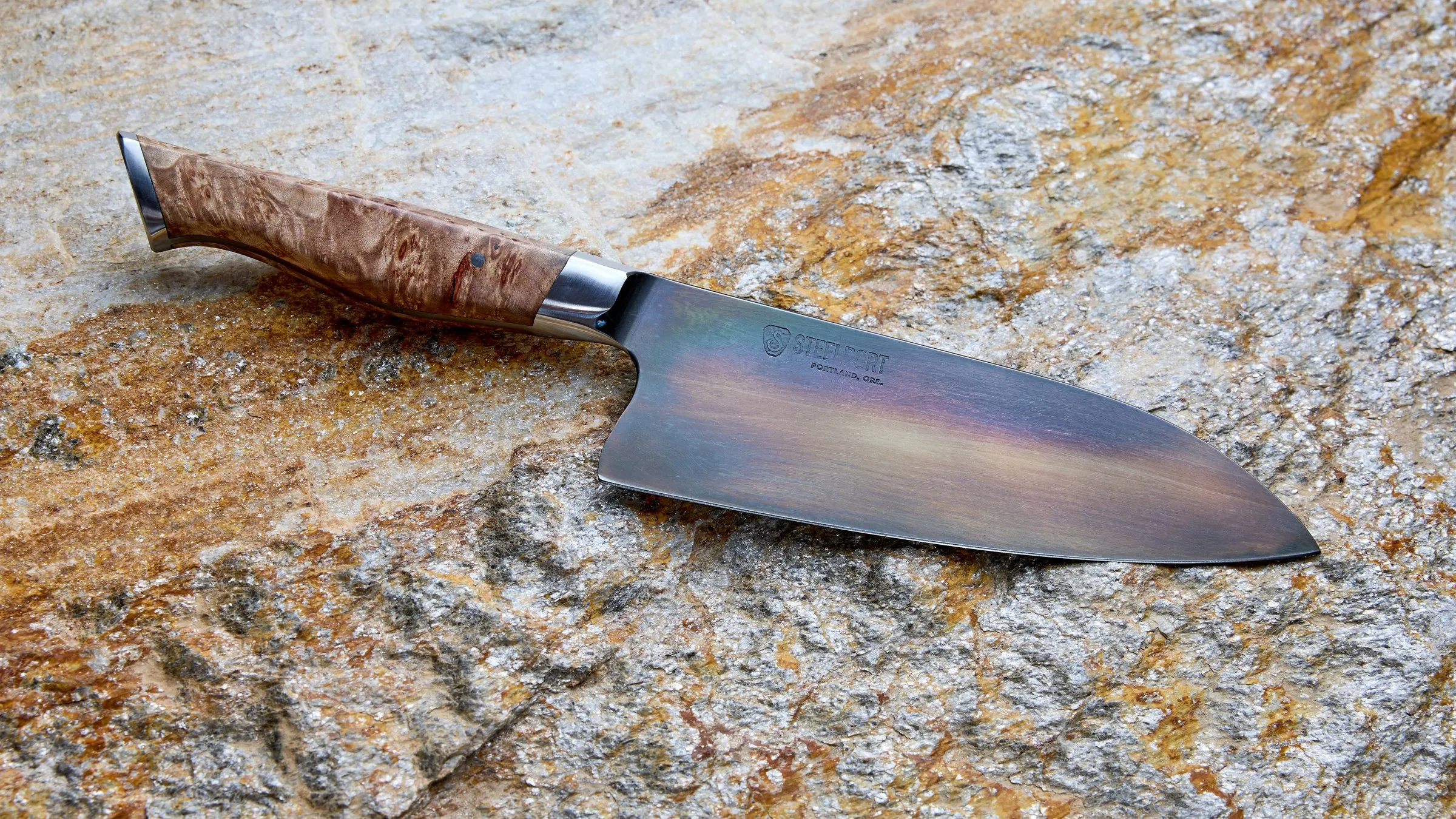 Faca de chef forjada à mão, de 15 cm, com a lâmina em tons de azul, roxo e dourado na superfície, cabo de madeira de raiz clara e virola de aço, sobre uma pedra cinza e ferrugem.