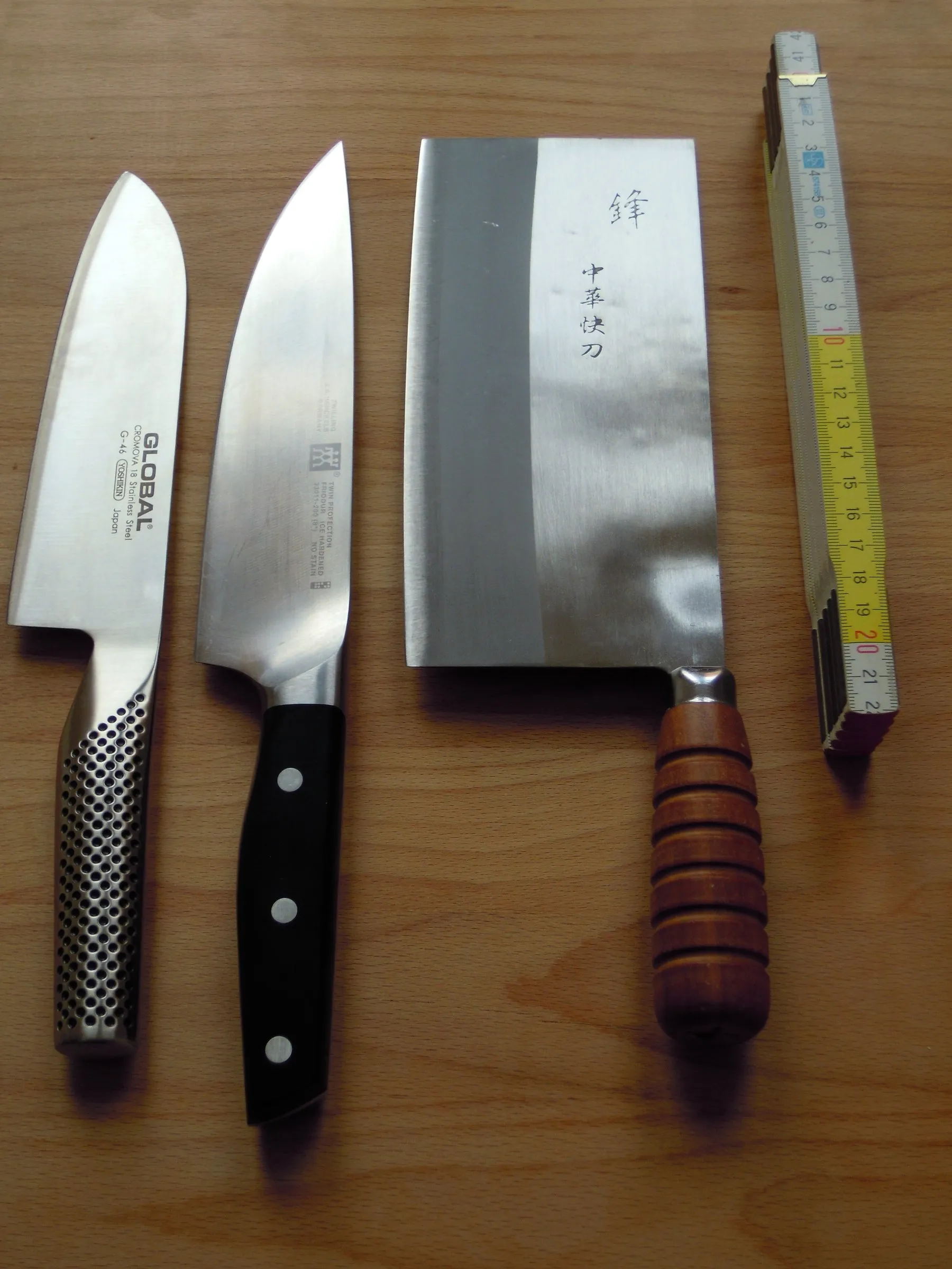 Três facas de cozinha lado a lado sobre uma mesa de madeira, com um metro dobrável ao lado: um santoku de aço inteiriço com cabo pontilhado, uma faca de chef alemã de cabo preto rebitado e uma faca chinesa retangular de cabo de madeira.