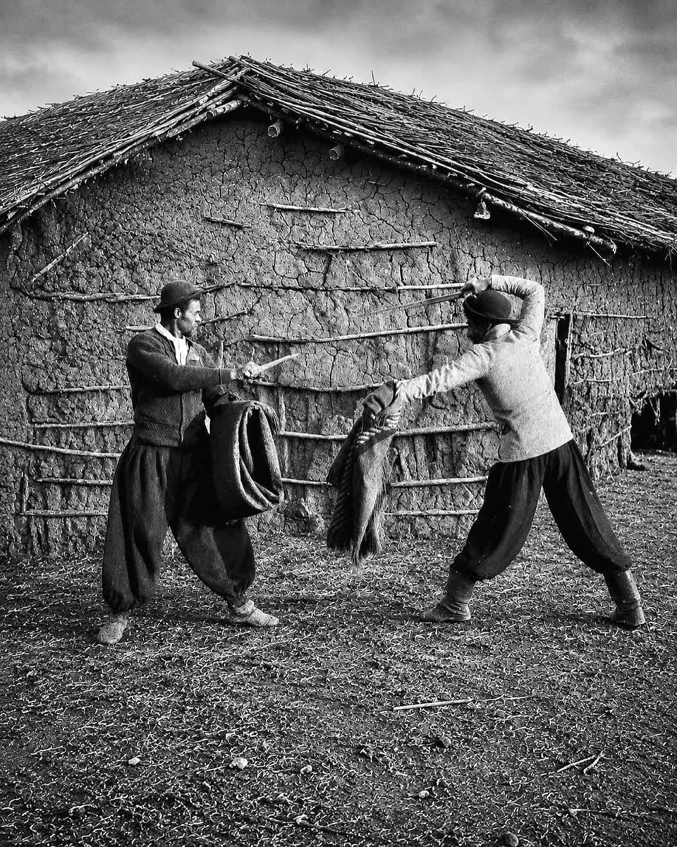 Fotografia de 1893 mostrando dois homens de bombacha e chapéu encenando um duelo de facas diante de um rancho de barro e palha, cada um com o poncho enrolado no braço como escudo