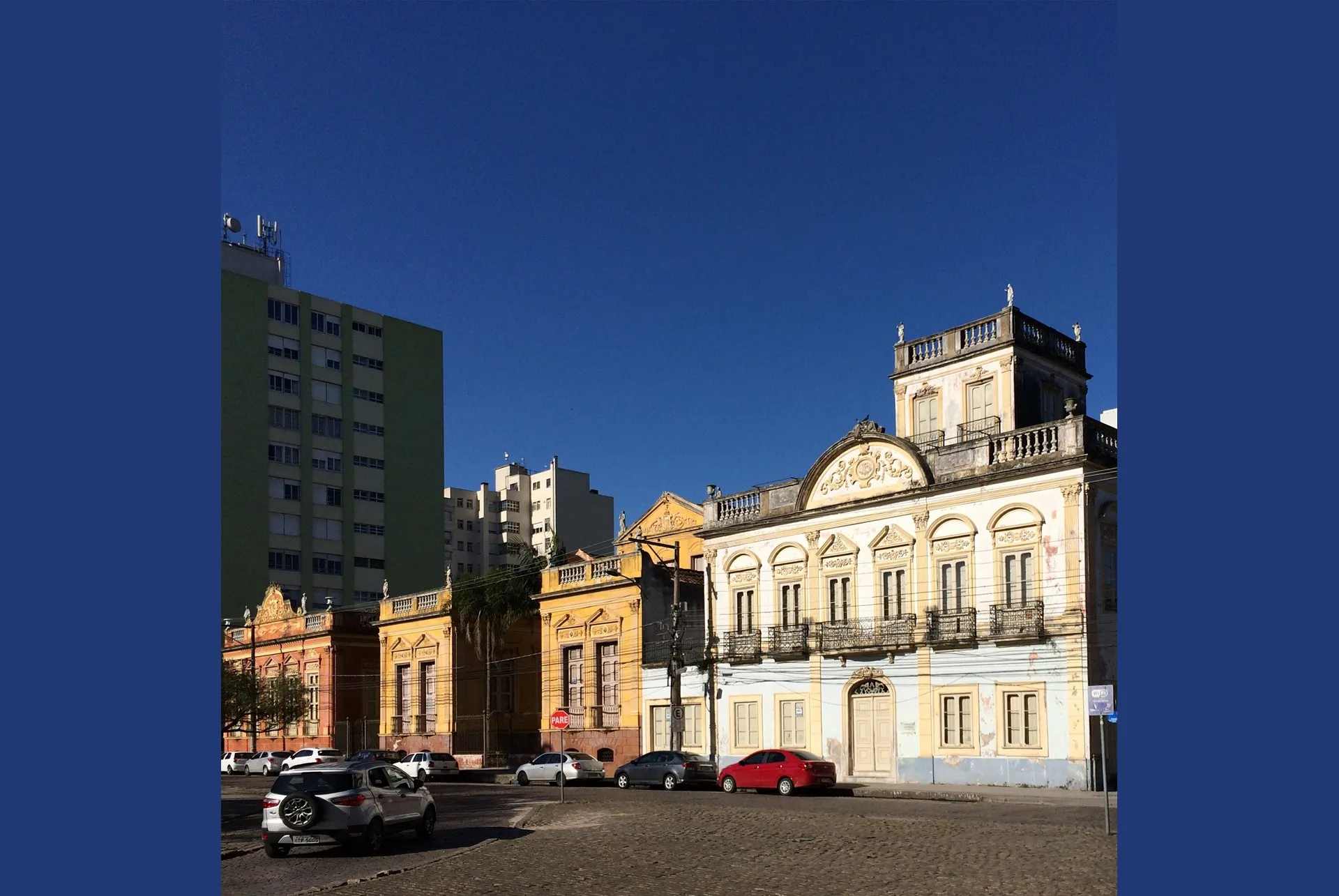 Fileira de casarões históricos de fachada eclética, em tons de amarelo, branco e vermelho, com sacadas de ferro trabalhado e platibandas ornamentadas, numa praça de pedras portuguesas de Pelotas, sob céu azul