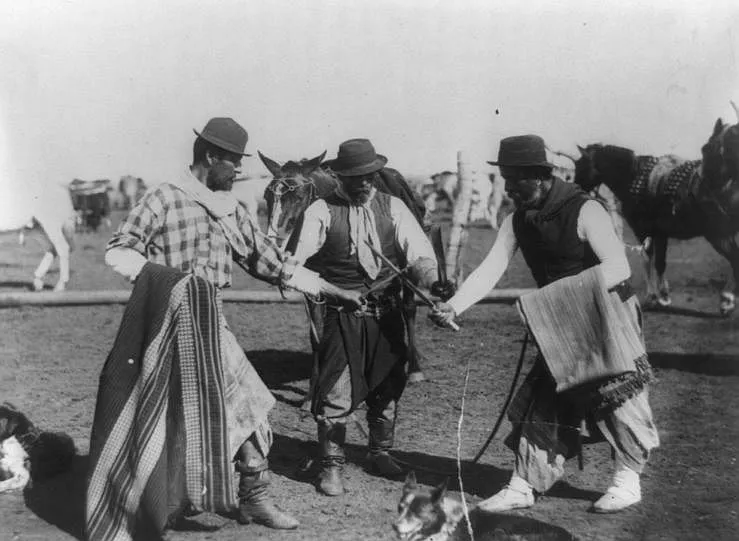Fotografia de cerca de 1890 mostrando três gaúchos no campo, dois deles cruzando facas enquanto um terceiro usa o poncho enrolado no braço como proteção, com cavalos ao fundo.