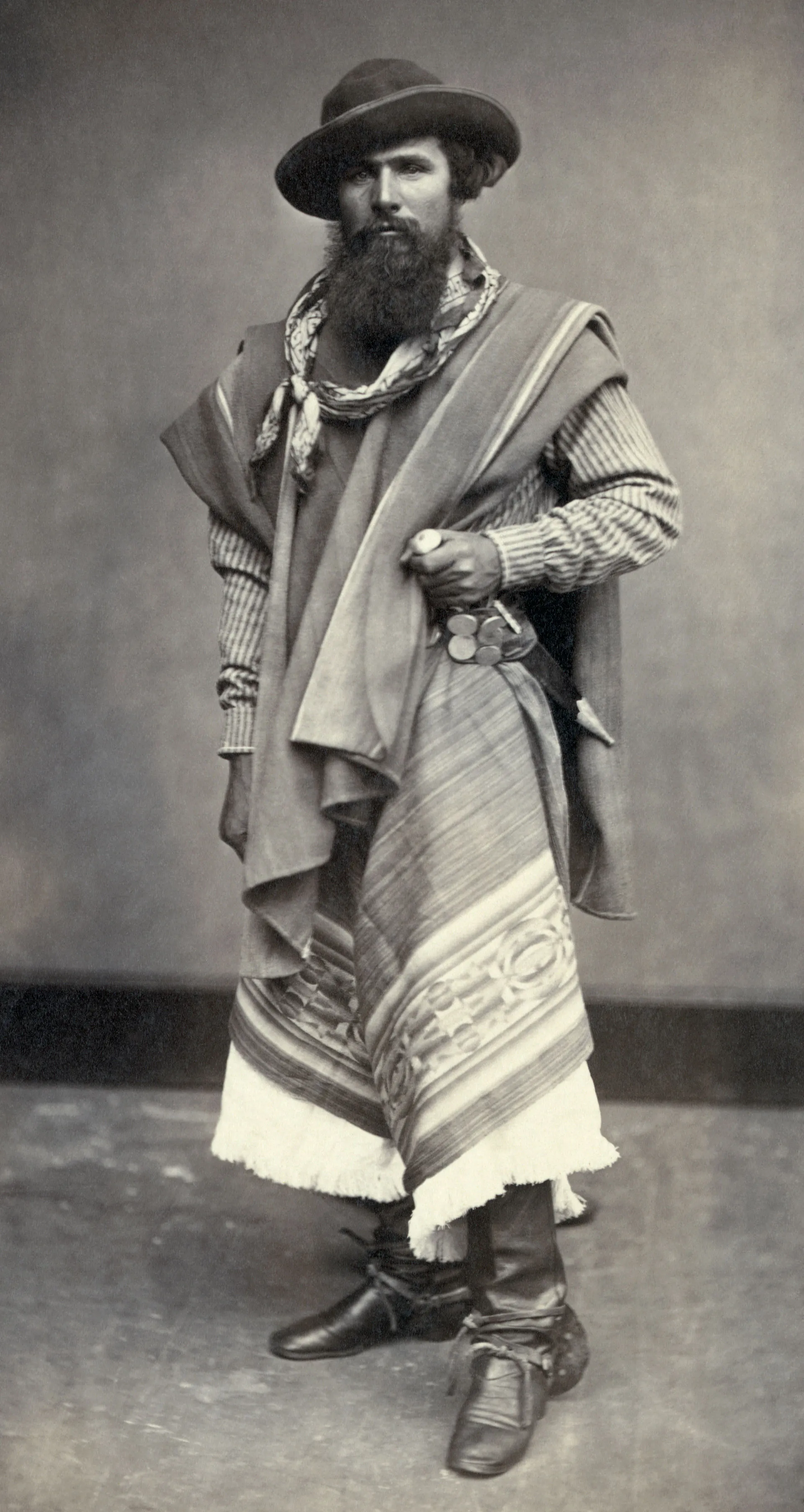 Retrato de estúdio de um gaúcho barbado de 1868, com chapéu, poncho e chiripá, tirador com rastra de moedas de prata na cintura e o cabo de um facão à mostra