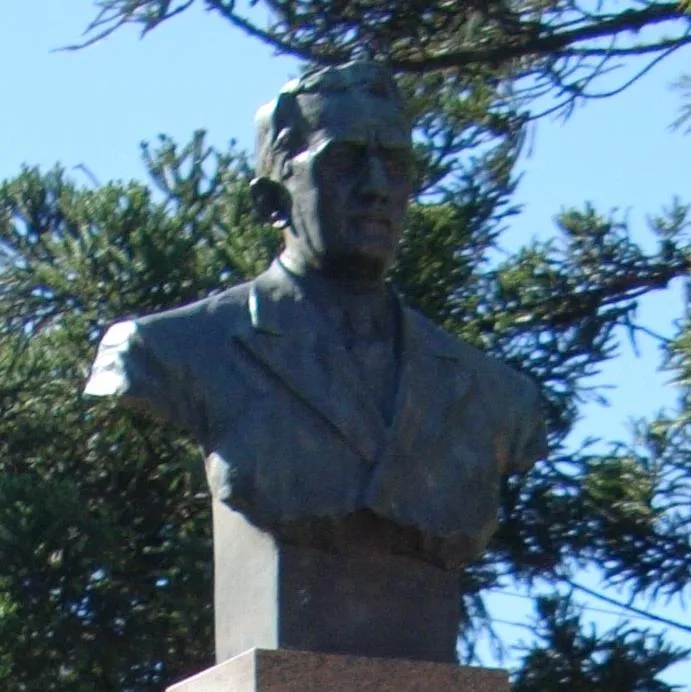 Busto de bronze de um homem de terno sobre pedestal de pedra, entre galhos de árvore