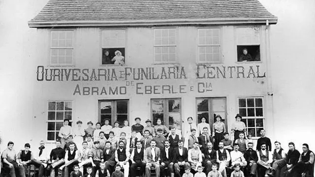 Fachada de sobrado com os dizeres "Ourivesaria e Funilaria Central — Abramo Eberle e Cia", com dezenas de operários, mulheres e crianças posando em frente, em 1907.