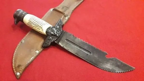 A mesma faca javali ao lado de sua bainha de couro costurada, com o serrilhado do dorso e o recorte característico junto à ponta bem visíveis