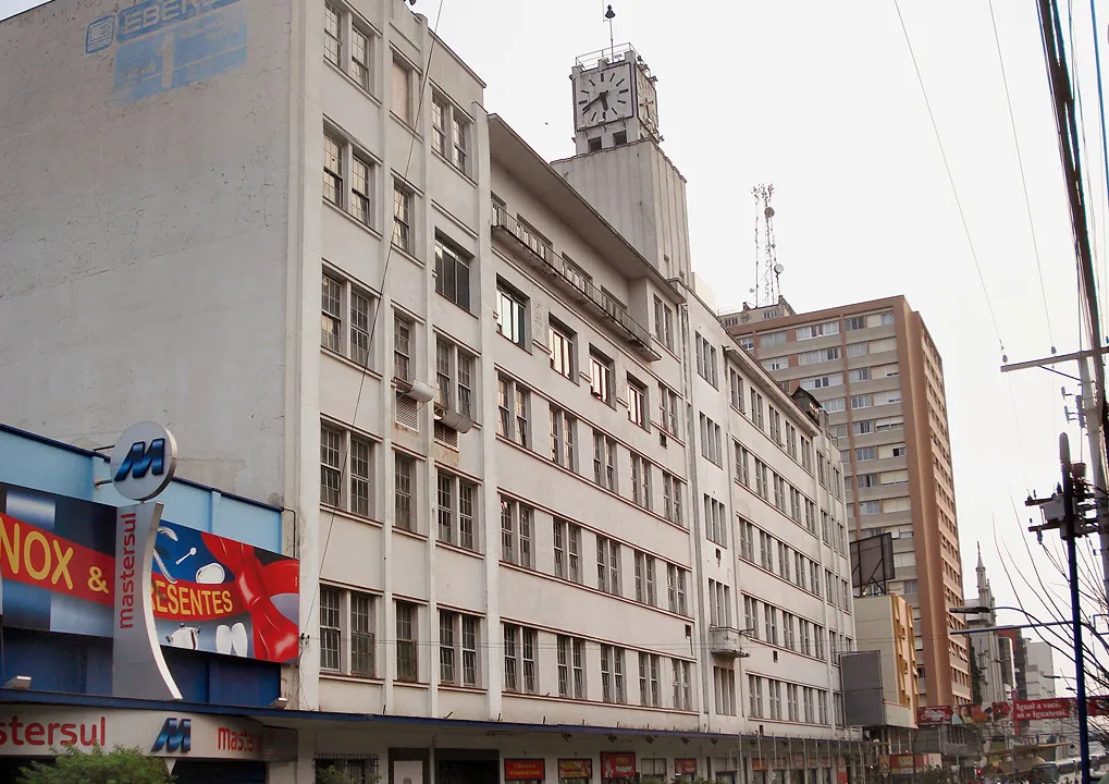 Prédio industrial branco de cinco andares com torre de relógio, o nome EBERLE ainda pintado na empena lateral, com lojas no térreo e movimento de rua, em Caxias do Sul.