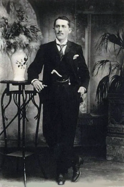 Retrato de estúdio de um homem jovem, de bigode, terno escuro e bengala, apoiado numa mesa de ferro