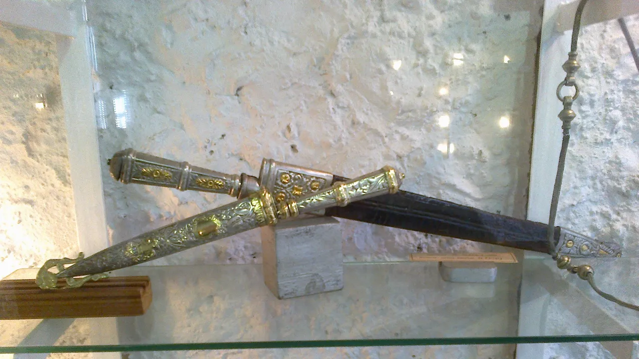 Dois cuchillos criollos com cabo e bainha de prata cinzelada e apliques de ouro, expostos cruzados numa vitrine de vidro contra uma parede de pedra caiada.