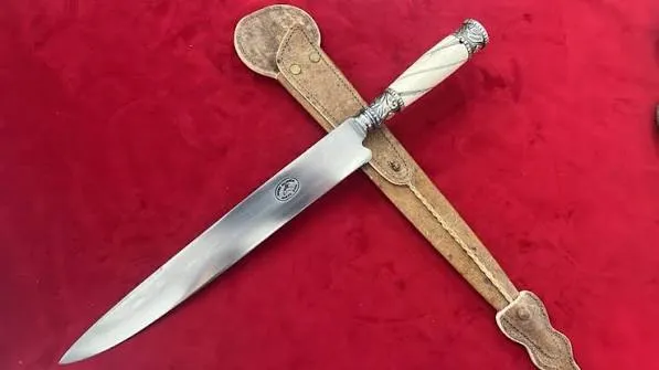 Cuchillo criollo Guazubirá com cabo de osso torneado em espiral e guarnições de alpaca, cruzado sobre a bainha de couro claro, com o carimbo oval visível na lâmina, sobre veludo vermelho