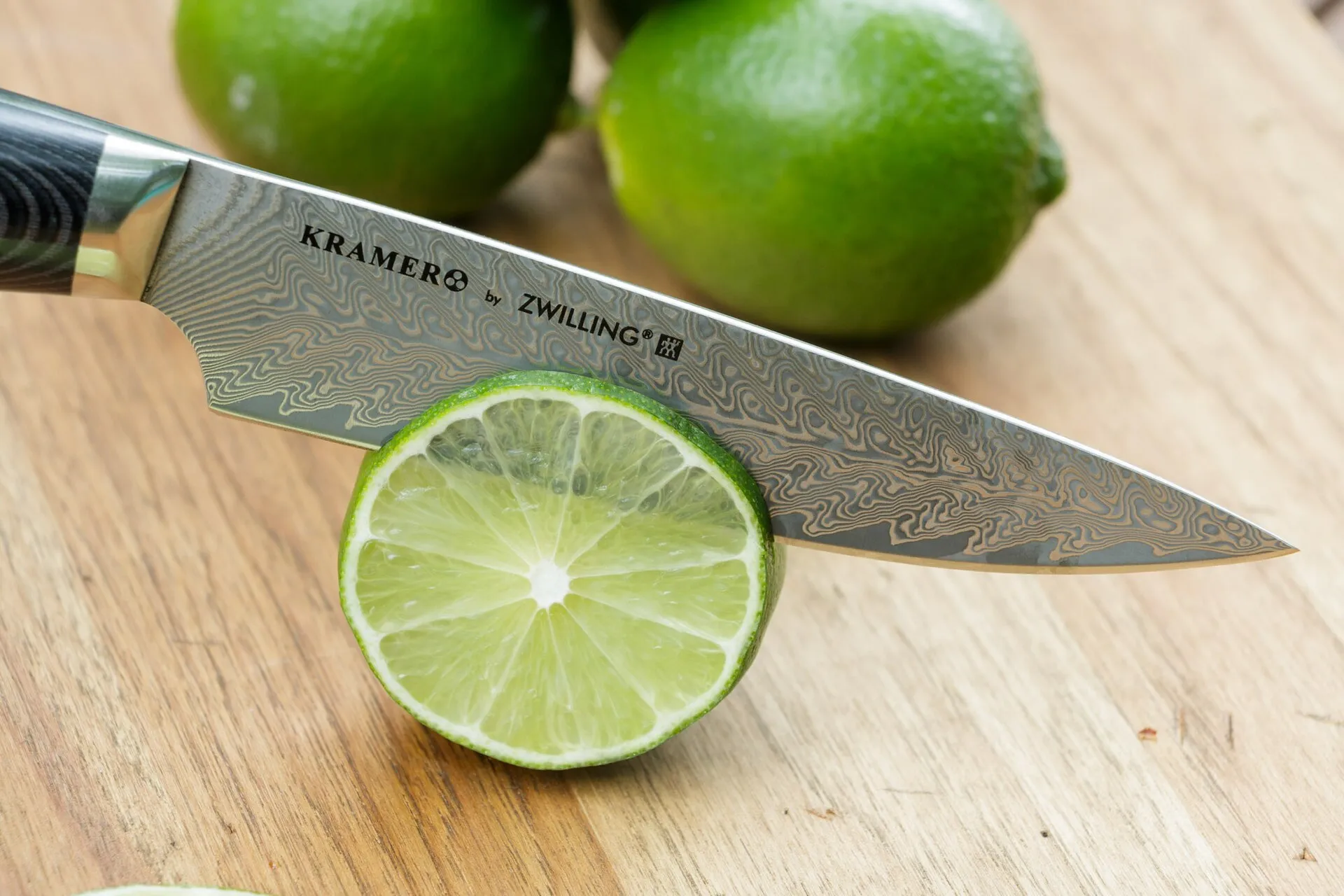 Faca de chef com lâmina de damasco marcada “KRAMER by ZWILLING”, cortando uma rodela de limão sobre tábua de madeira