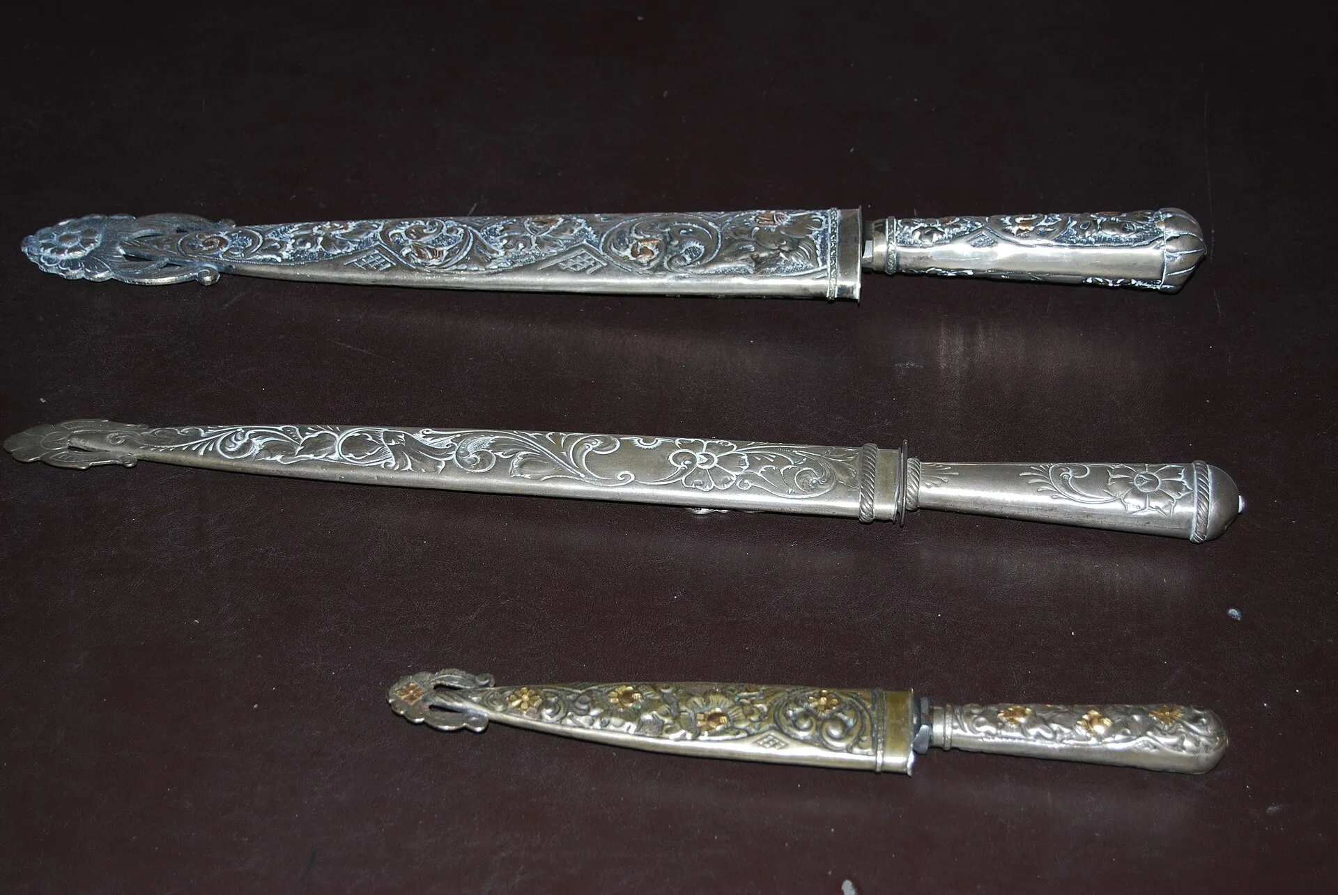 Três facones criollos com bainha e cabo de prata trabalhada, em três tamanhos, dispostos sobre fundo escuro; a prataria mostra motivos florais cinzelados