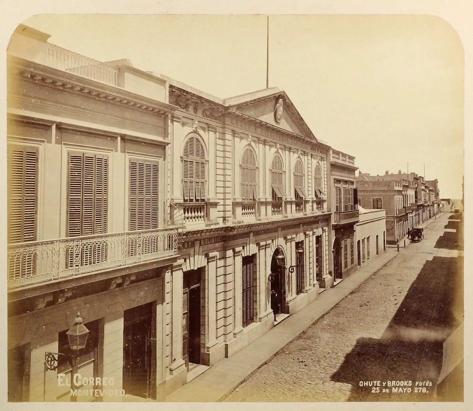 Fotografia de 1873 de uma rua da Cidade Velha de Montevidéu, com sobrados de sacadas de ferro, o prédio do Museo y Biblioteca Nacional e a placa de El Correo na esquina
