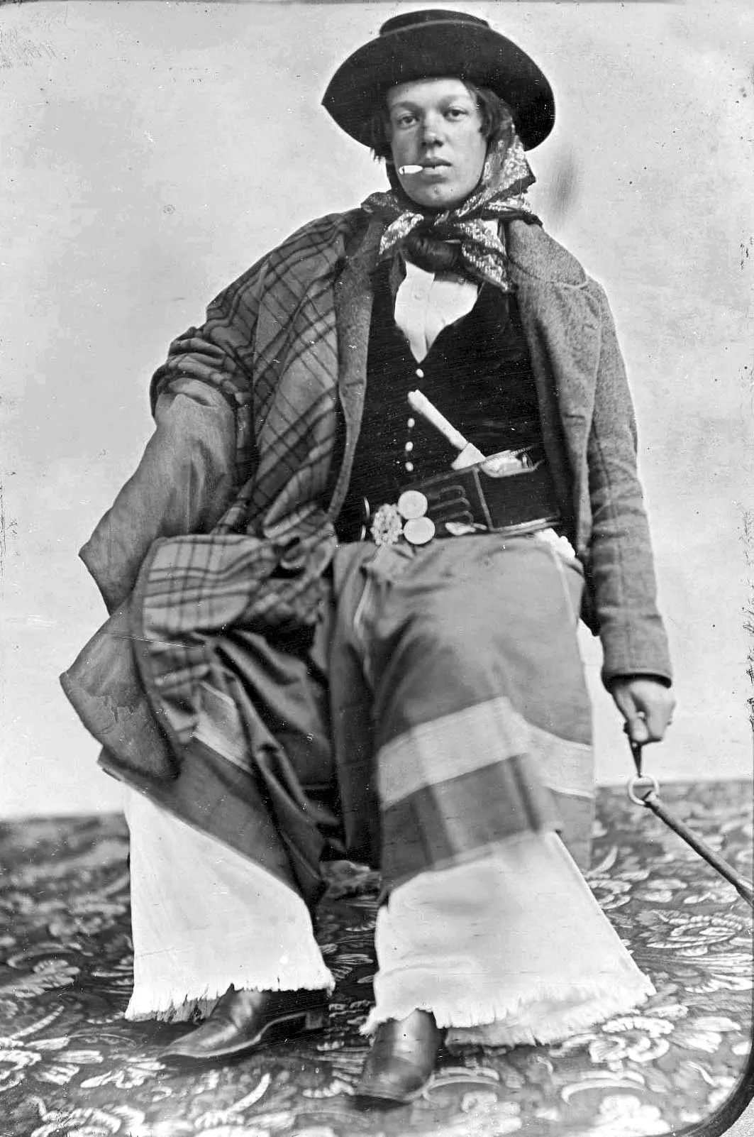Fotografia em preto e branco do século XIX de um gaúcho sentado, de poncho, lenço e bombachas, com o cabo da faca aparecendo na cintura sobre o tirador enfeitado de moedas