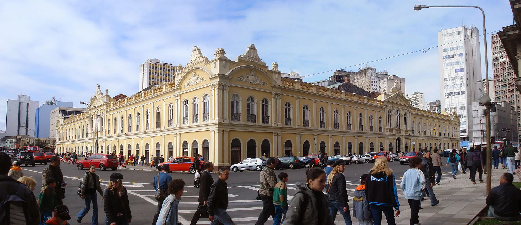 Fachada de um casarão eclético amarelo de dois andares, com pátio movimentado de gente e carros em primeiro plano e prédios modernos ao fundo