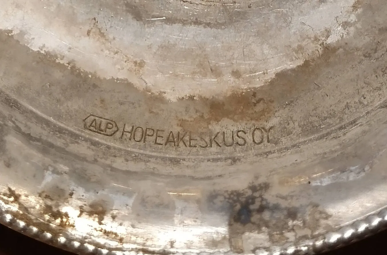 Macro de um carimbo gravado no fundo de uma peça metálica manchada: a sigla ALP dentro de um losango, seguida do nome HOPEAKESKUS OY