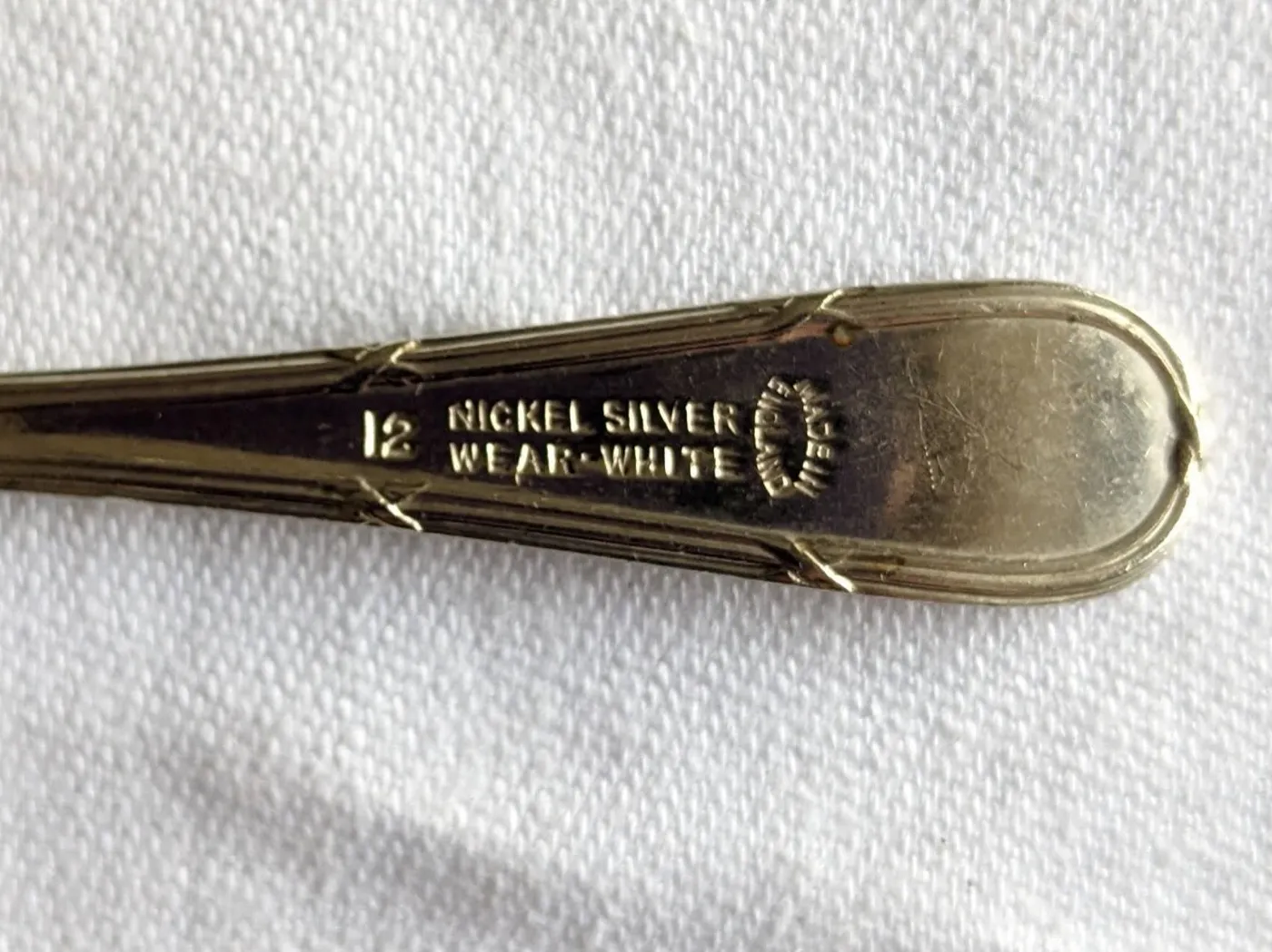 Cabo de colher de tom amarelado marcado com o número 12 e as palavras NICKEL SILVER WEAR WHITE, ao lado de um selo oval com os dizeres MADE IN ENGLAND