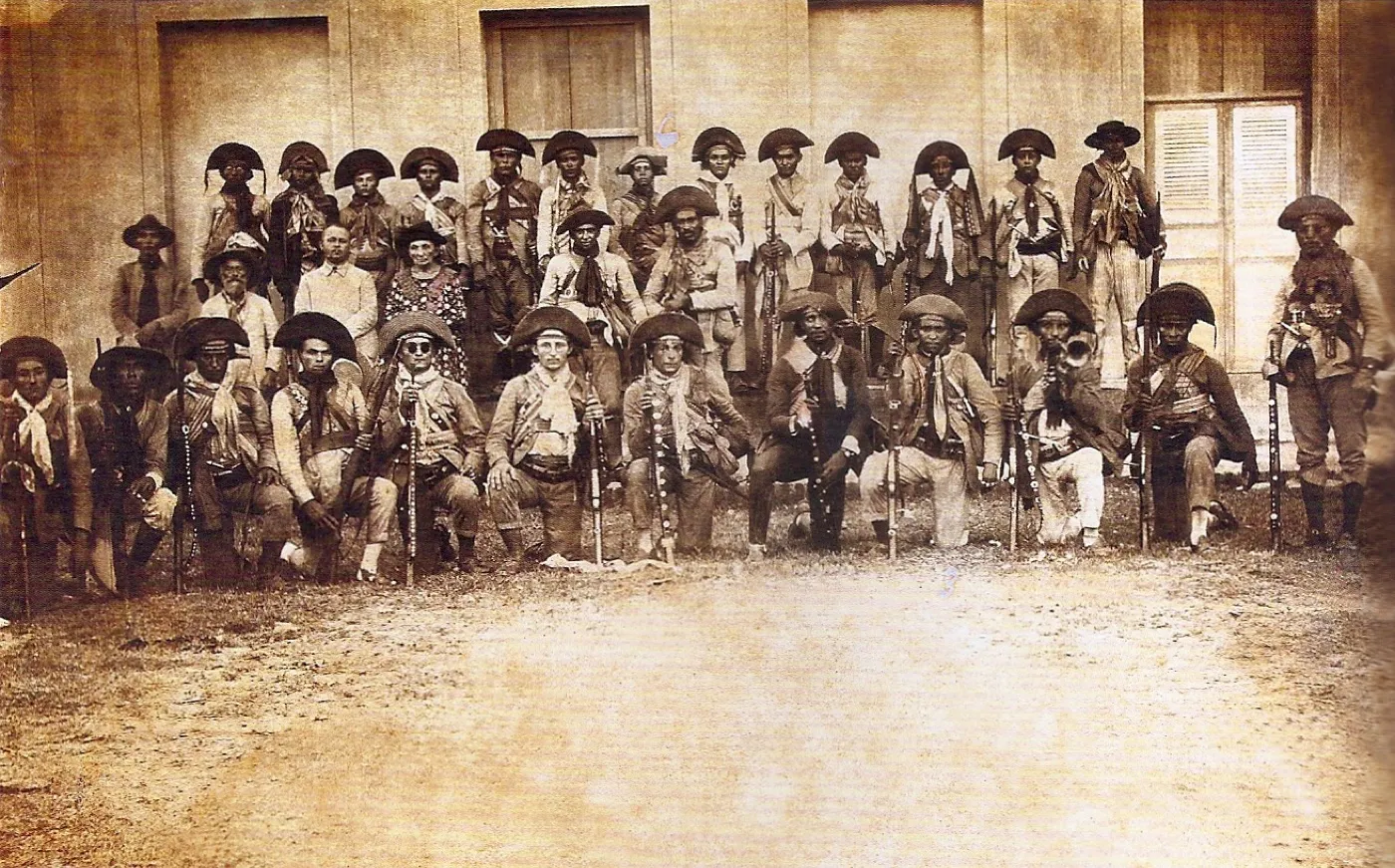 Fotografia em preto e branco de 1927 do bando de Lampião enfileirado diante de um casarão em Limoeiro do Norte, Ceará, cerca de trinta cangaceiros de chapéu de couro, lenço e cartucheiras cruzadas no peito, portando rifles, facas e punhais na cintura