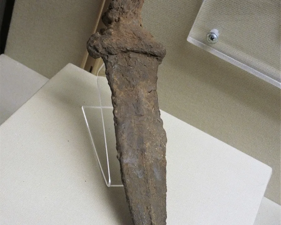 Fotografia de um pugio romano de ferro, dos séculos I e II d.C., com lâmina larga triangular de dois gumes corroída pelo tempo e cabo curto em forma de cruz, exposto em pé sobre um suporte de acrílico transparente numa vitrine de museu.