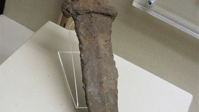 Fotografia de um pugio romano de ferro, dos séculos I e II d.C., com lâmina larga triangular de dois gumes corroída pelo tempo e cabo curto em forma de cruz, exposto em pé sobre um suporte de acrílico transparente numa vitrine de museu.