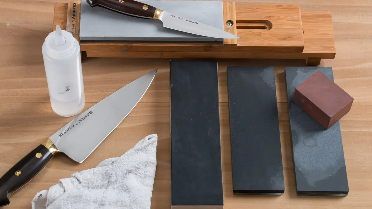 Bancada de afiação com duas facas de cozinha, um suporte de bambu e quatro pedras de granulações diferentes alinhadas sobre a madeira, ao lado de um borrifador e um pano.