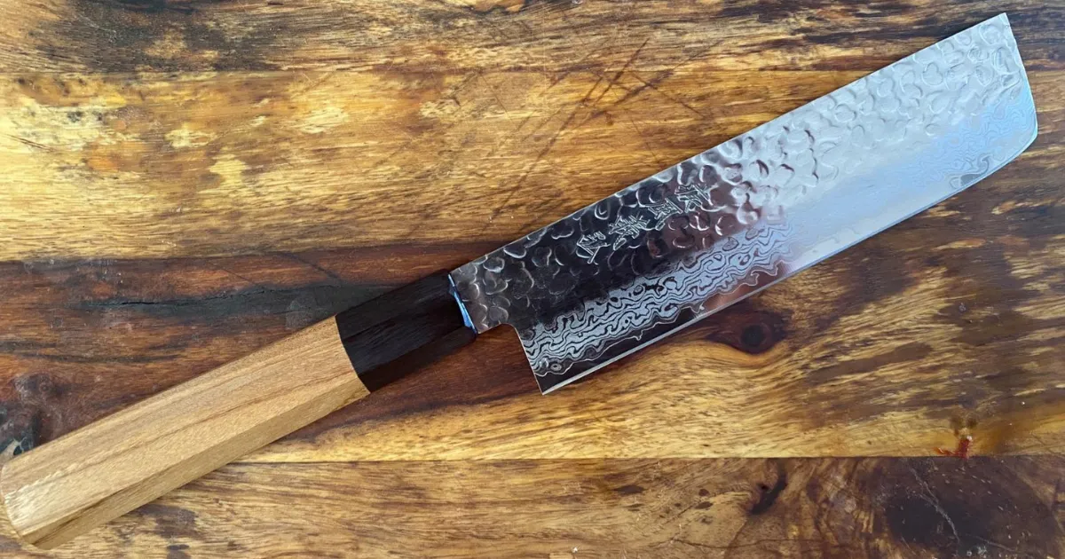 Faca japonesa nakiri com acabamento martelado, lâmina mostrando linha ondulada entre o núcleo e a capa, cabo de madeira clara