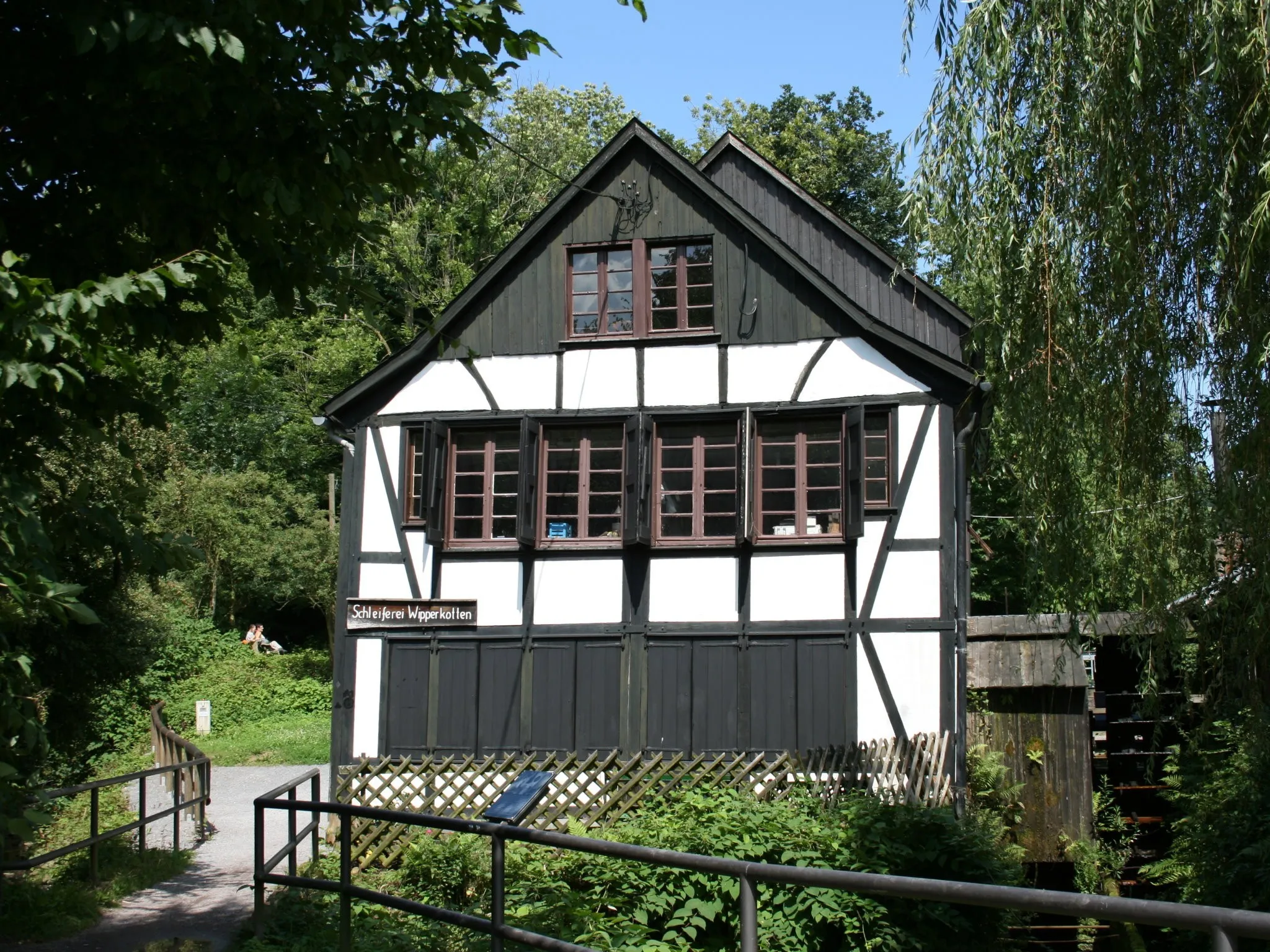 Casa de enxaimel branca e preta à beira de um curso d’água, com placa “Schleiferei Wipperkotten” e uma roda d’água de madeira ao lado