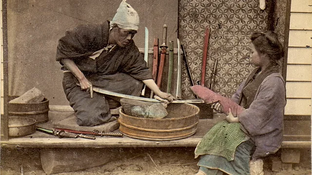 Fotografia colorida à mão, de cerca de 1909: um afiador japonês ajoelhado passa uma espada sobre uma pedra apoiada numa tina de madeira, com outras espadas encostadas na parede atrás dele e uma mulher sentada ao lado segurando uma lâmina embainhada.