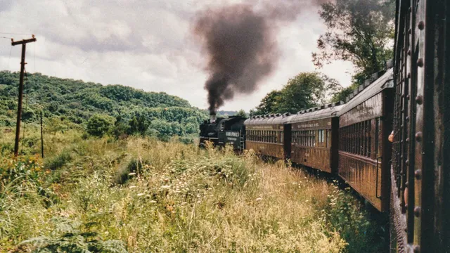 Locomotiva a vapor "Maria Fumaça" puxando vagões de madeira escura pela Serra Gaúcha, soltando fumaça preta contra um céu nublado, cercada de mato alto e vegetação.