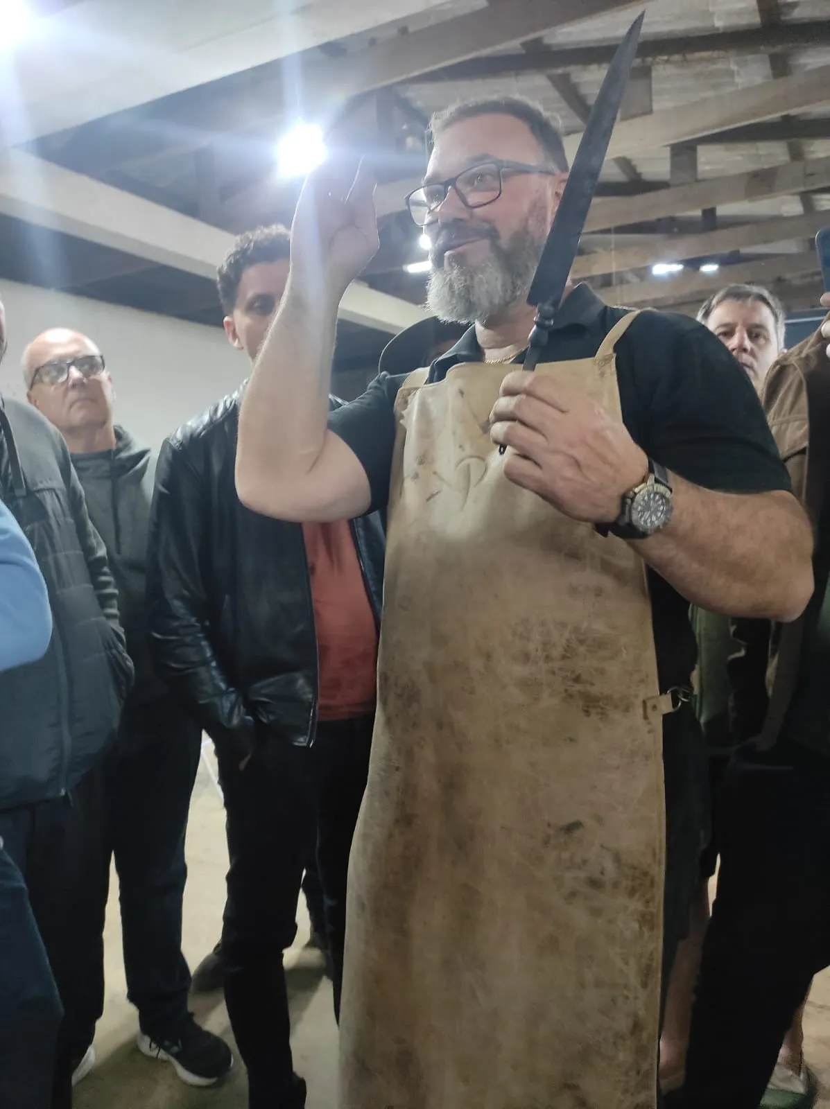 Sandro Boeck, cercado por participantes, ergue uma lâmina de damasco escurecida pelo ácido e aponta para ela com a outra mão, enquanto explica.