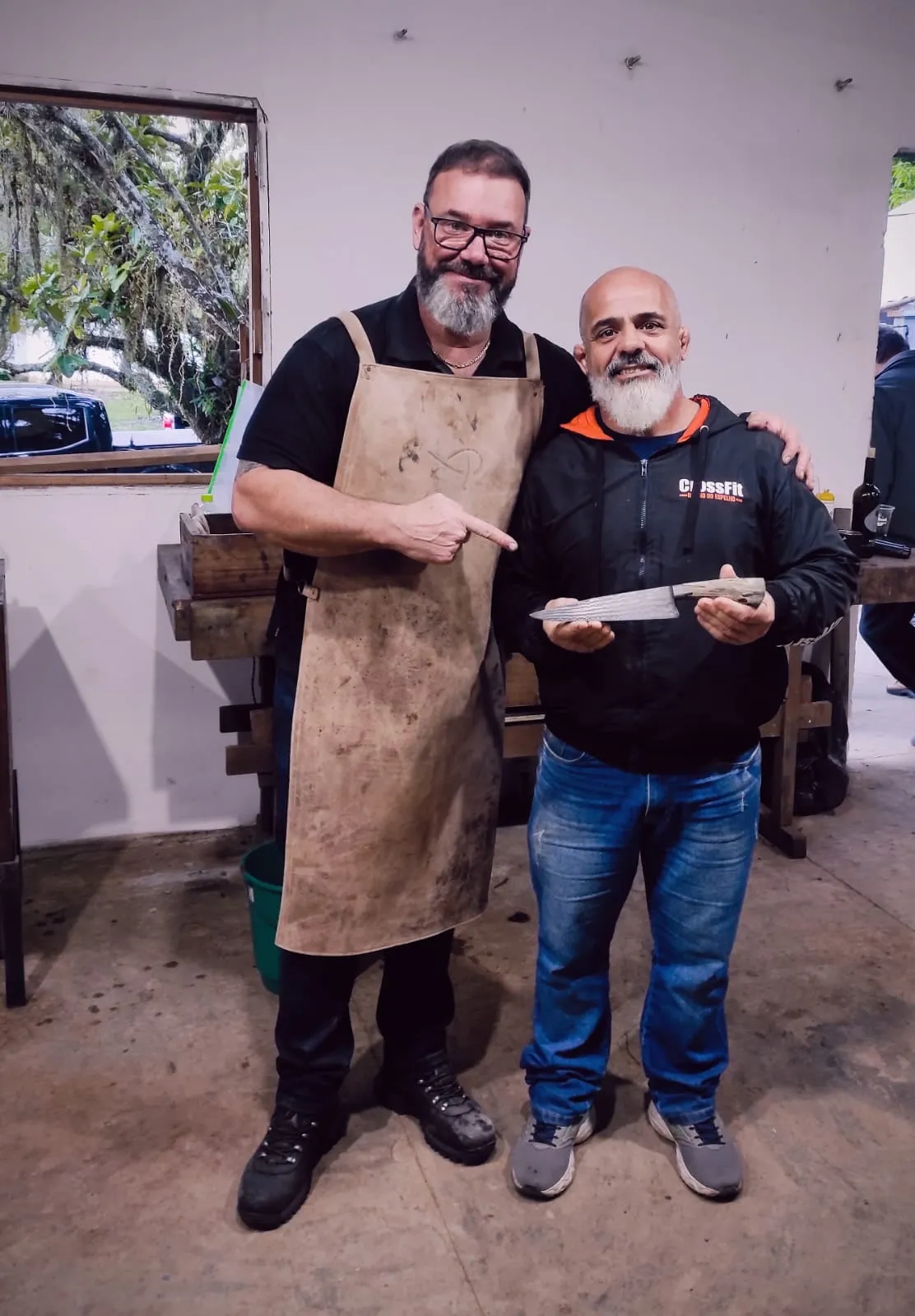 Sandro Boeck, de avental de couro, aponta sorrindo para Walter Fonseca, que segura com as duas mãos uma faca de damasco com cabo claro, em pé diante da bancada da oficina.