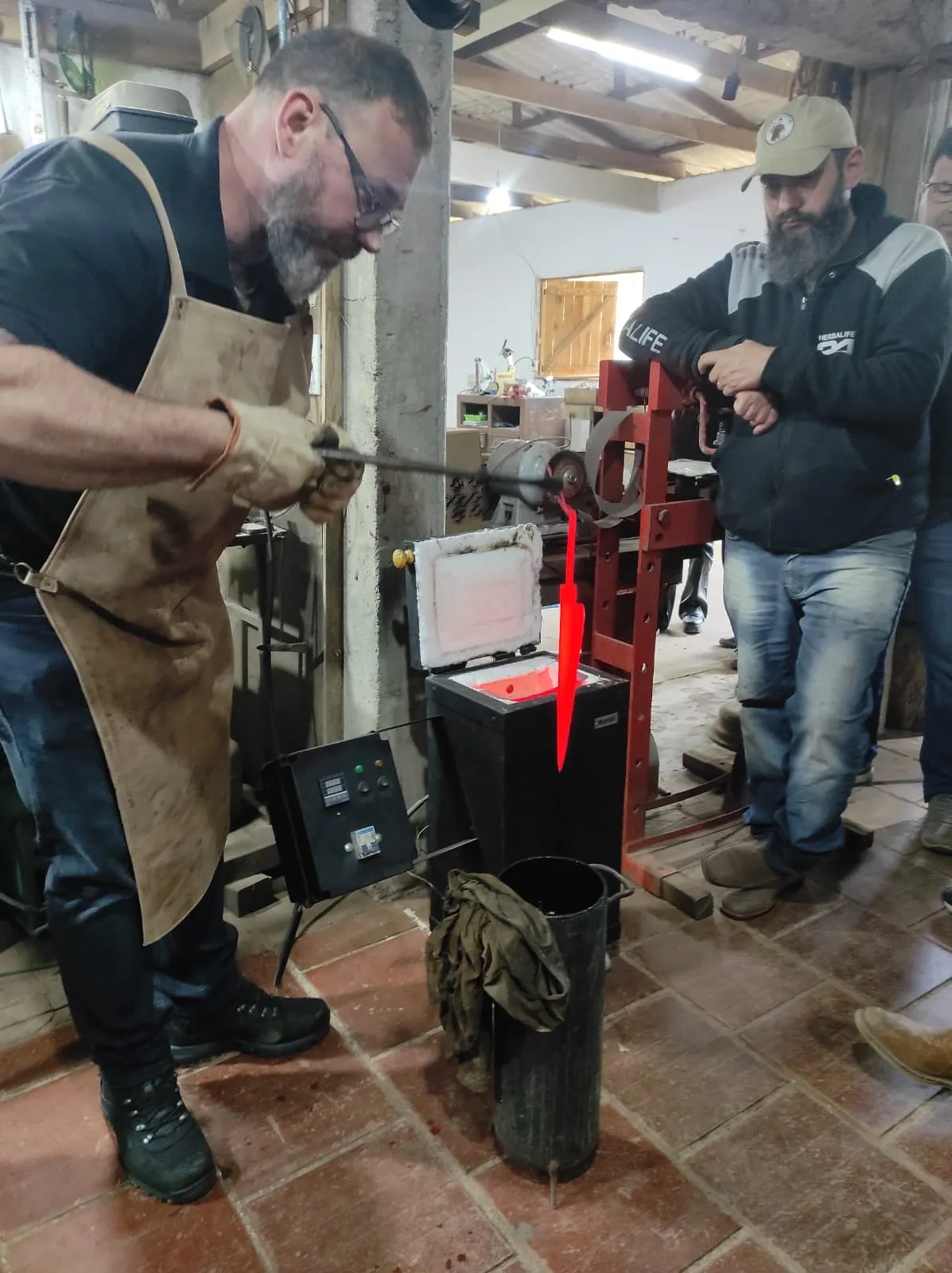 Sandro Boeck, de avental de couro e luvas, retira do forno uma lâmina incandescente, vermelha da ponta à espiga, enquanto dois participantes observam a poucos passos.