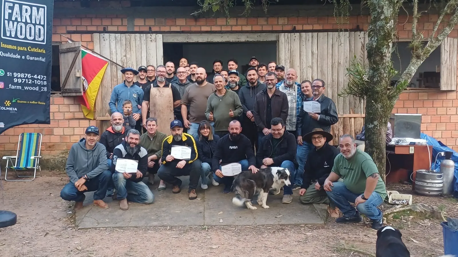 Foto de grupo da 12ª Vivência de Cutelaria: mais de trinta participantes em pé e agachados diante do galpão de tijolos, alguns segurando certificados, com Sandro Boeck de avental ao centro, a bandeira do Rio Grande do Sul numa janela e um cão border collie na frente.